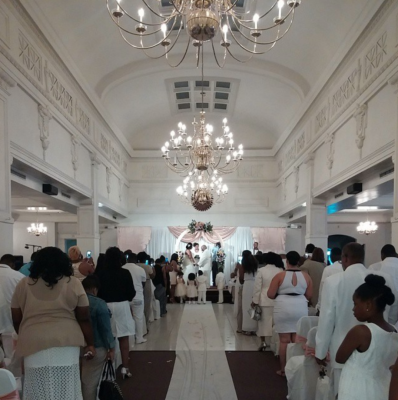 A newlywed couple shares their first dance in a grand, historic reception hall with high ceilings, ornate chandeliers,